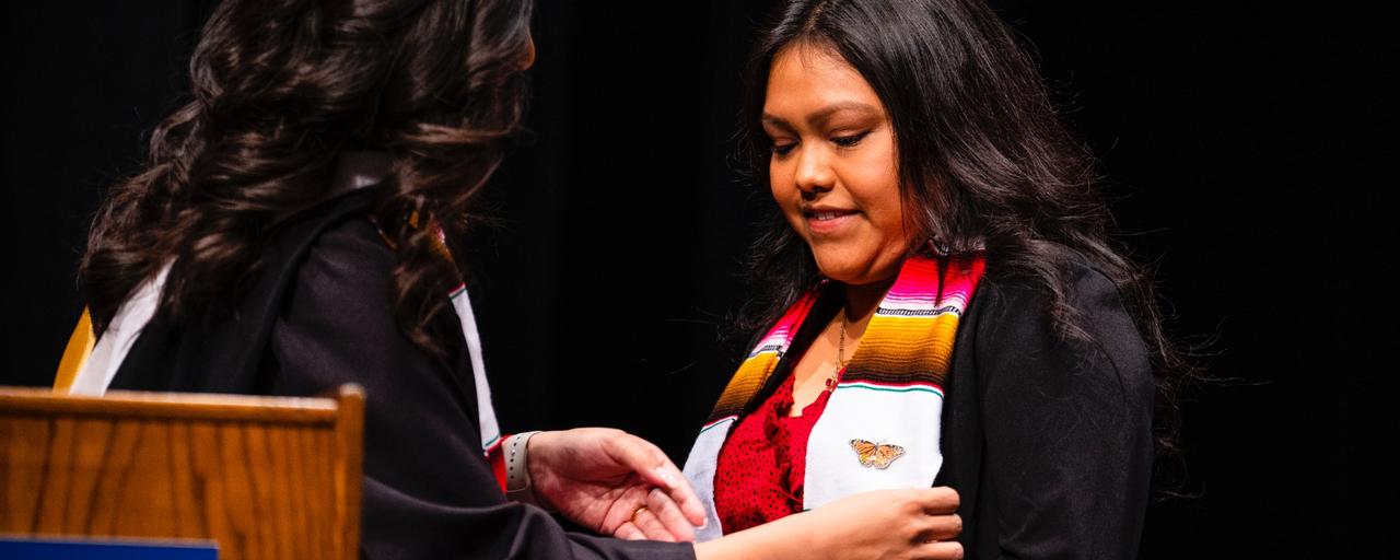 Student in graduation regalia being pinned with a monarch butterfly pin